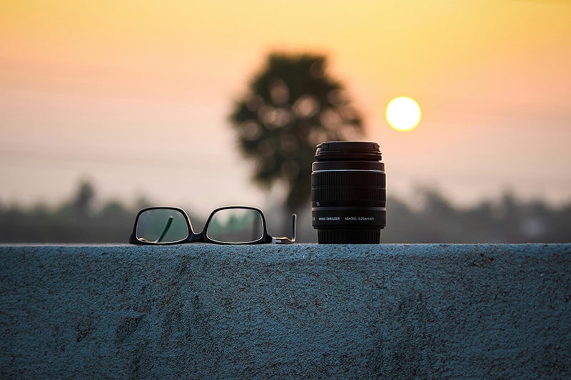 A picture showing a zoom photography lens next to a pair of viewing glasses with a sunset as the background.