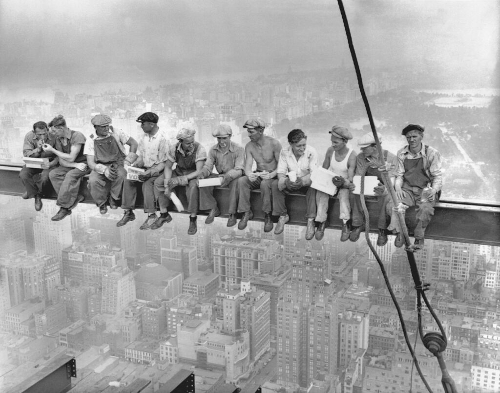 Eleven ironworkers sitting in a row on a steel girder high above a city, with their feet dangling. They are eating lunch, talking, and smoking, seemingly unconcerned by the extreme height.