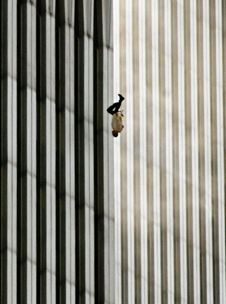 A person in dark trousers and a white top falls vertically, headfirst, against the vertical lines of the World Trade Center towers.