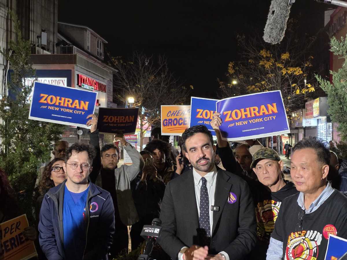 Zohran Mamdani speaking at a podium during a press conference at Diversity Plaza in Jackson Heights, with microphones in front and supporters in the background