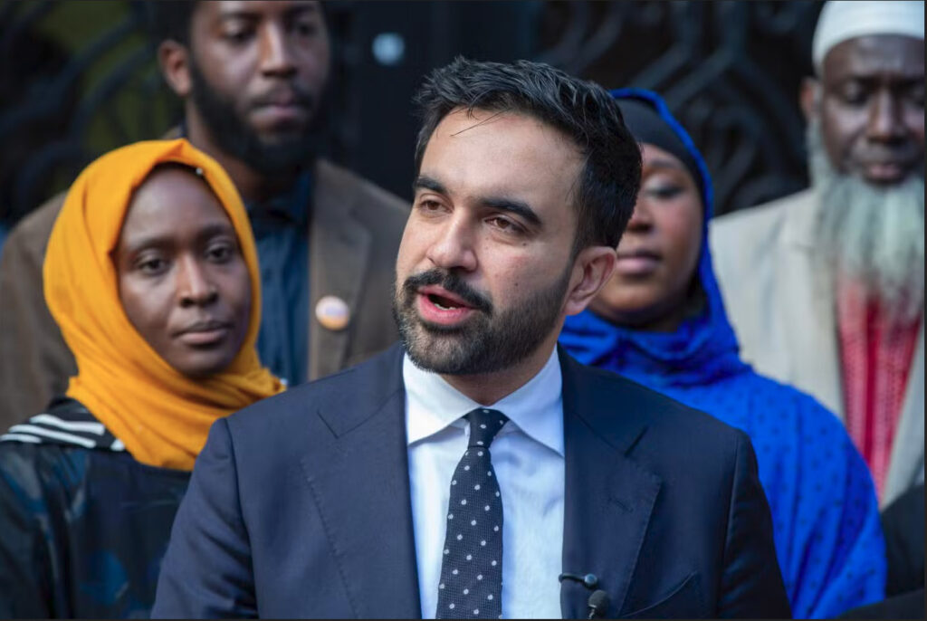 Zohran Mamdani standing at a microphone outside a mosque in the Bronx, visibly moved and speaking to reporters, with faith‑leaders behind him and a serious expression on his face.