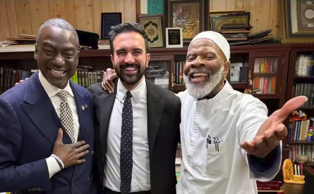 Zohran Mamdani, Yusef Abdus Salaam, and Imam Siraj Wahhaj standing together outside a Brooklyn mosque, smiling during a public meeting with community members.