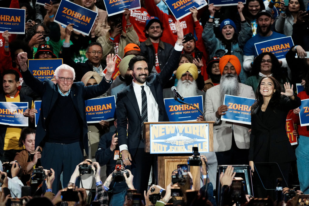 Zohran Mamdani, Bernie Sanders, and Alexandria Ocasio-Cortez standing on stage, smiling and waving to supporters at the “New York is Not For Sale” rally in Forest Hills Stadium, Queens, October 2025.