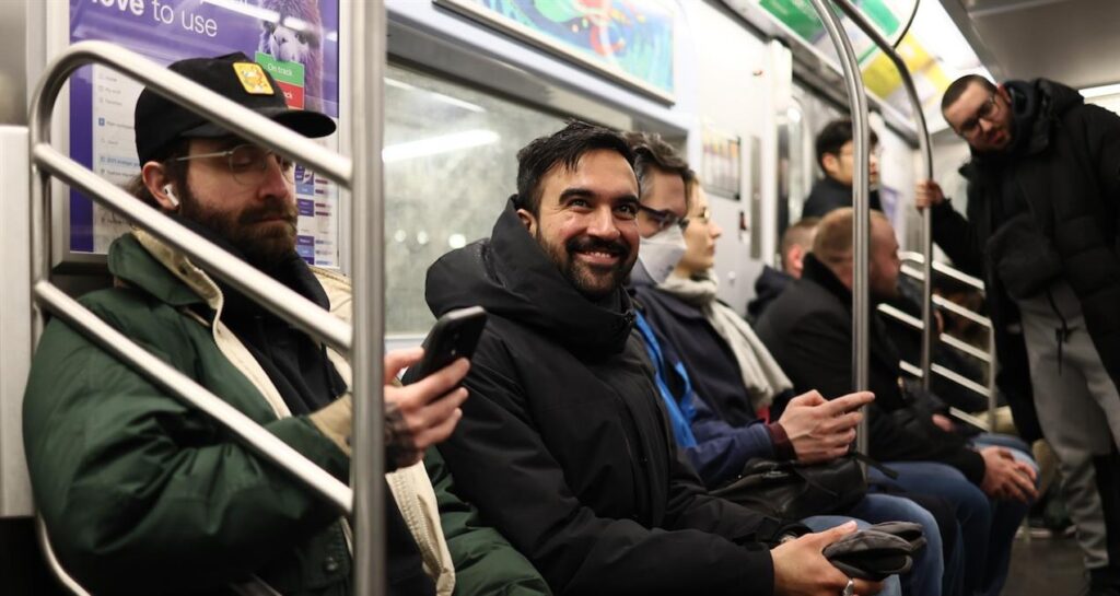 Zohran Mamdani seated on a subway train, smiling and holding a phone, surrounded by campaign team members in a candid transit moment.