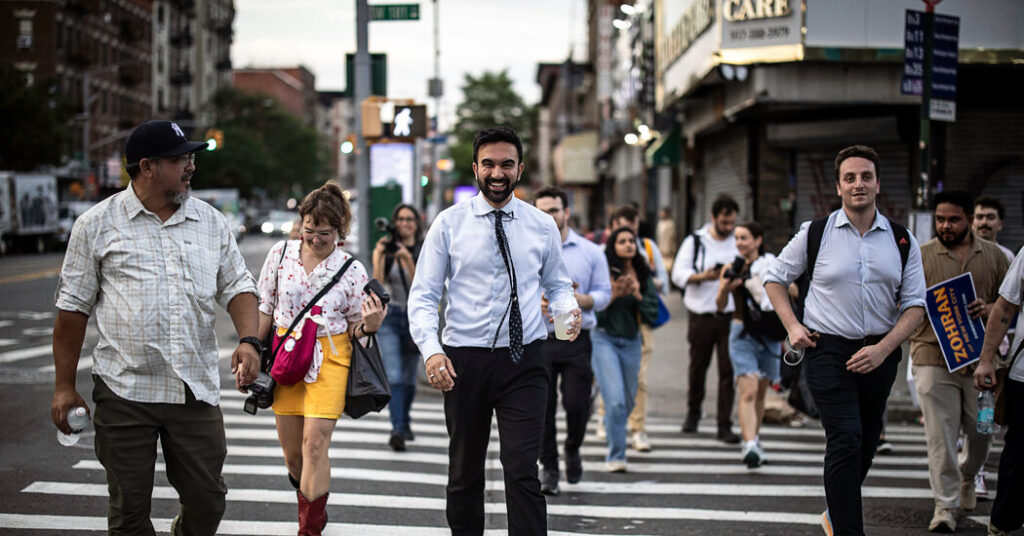 Zohran Mamdani smiling as he crosses a Manhattan street, surrounded by pedestrians, during a walking tour of the city in the final days of his mayoral campaign.