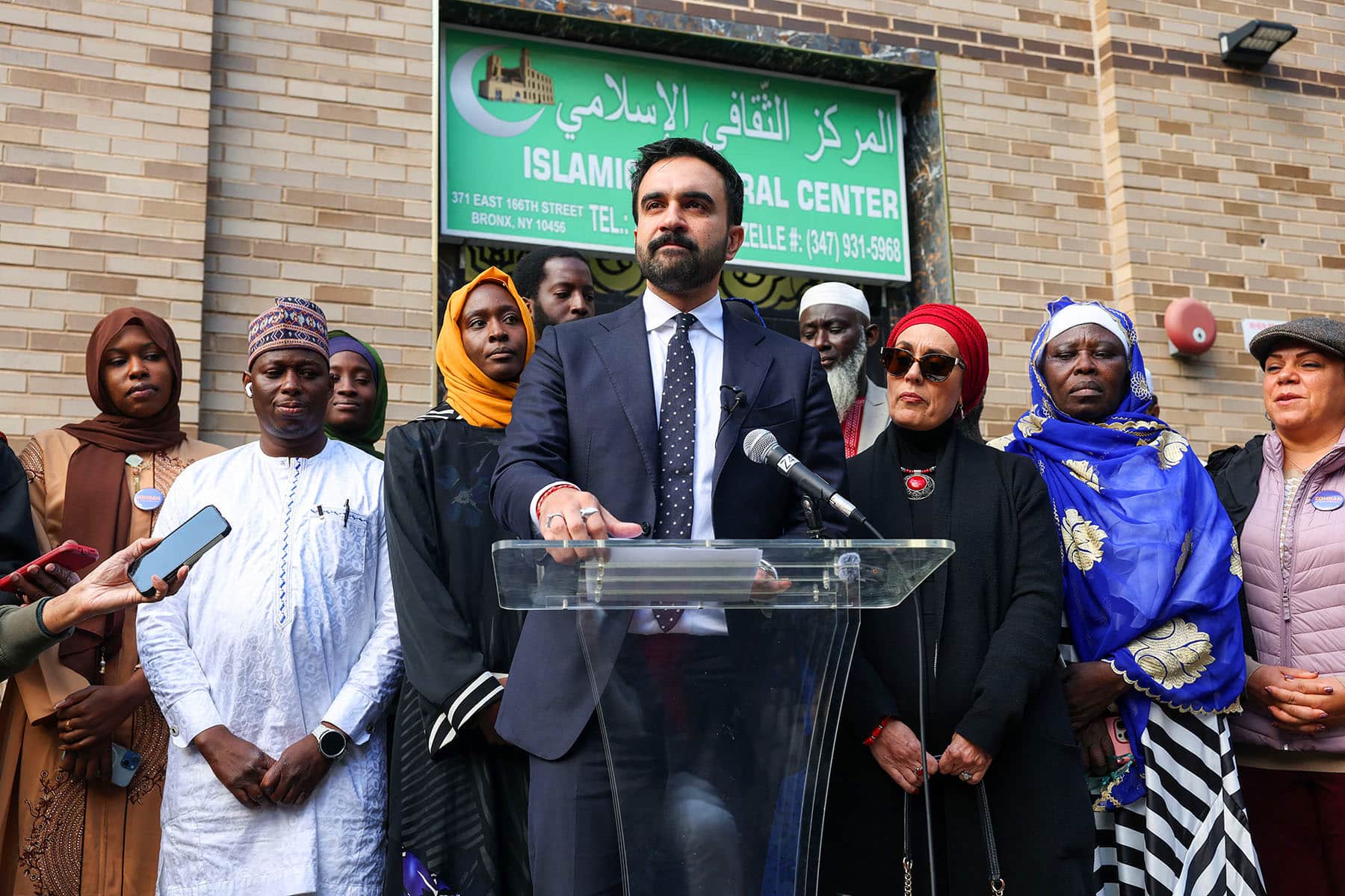 Zohran Mamdani stands in front of a microphone outside a mosque in the Bronx, his expression resolute as he engages with reporters about Islamophobia in his mayoral campaign.